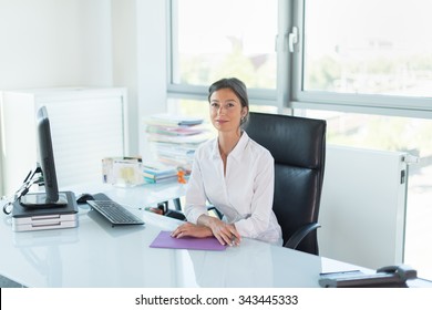 Assistant Manager Looking At Camera In Her White Office Woman Sitting At A Tidy Glass Desk In Front Of A Black Computer She Is Wearing A White Shirt Her Hair Are Tied Her Hands Are Crossed On A Folder