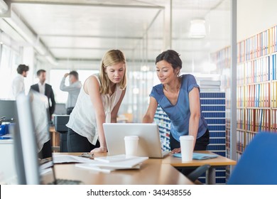 Assistant And Partner Coffee Meeting. Focus On Two Women In Her 30s And 40s Who Are Standing In Front Of A Laptop And Two White Coffee Cup, In A Luminous Open Space Looking At Balance Sheets