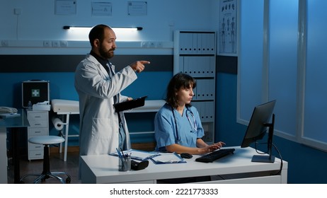 Assistant Standing In Front Of Pc Showing Medical Expertise To Medic Discussing Patient Illness Diagnosis Planning Together Health Care Treatment. Staff Working Late At Night In Hospital Office