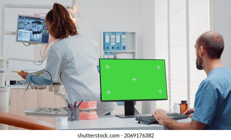 Assistant Using Horizontal Green Screen On Computer While Dentist Preparing Equipment For Oral Care Examination In Dental Cabinet. Man Looking At Chroma Key And Mockup Template On Monitor