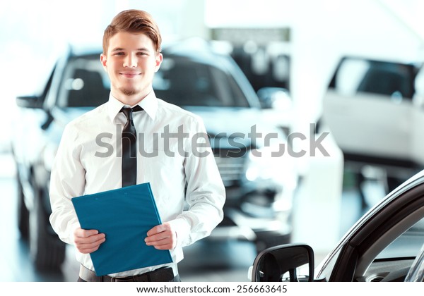 Assistant in vehicle search. Portrait of a\
handsome young car sales man in formalwear holding a clipboard and\
looking at camera in a car dealership\
