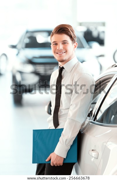 Assistant in vehicle search. Portrait of a\
handsome young car sales man in formalwear holding a clipboard and\
looking at camera in a car dealership\
