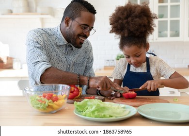 Assisting Daddy. Happy Smiling Single African Black Dad And Small Tween Daughter Engaged In Easy Cooking Together Spending Time At Kitchen Talking Preparing Healthy Vegan Lunch Of Lettuce And Pepper