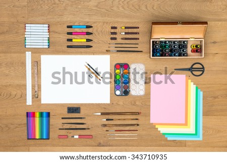Assorted array of arts, crafts, drawing and design items, such as pens, rulers, markers, felt tip pens, fountain pens brushes, watercolor, and various sheets of paper on a wooden surface