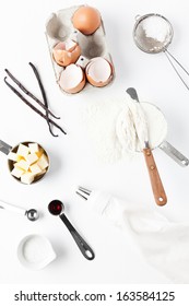 Assorted Baking Ingredients And Tools: Eggshells, Vanilla Pods, Flour In Measuring Cup, Spatula, Measuring Spoon With Vanilla Extract, Butter, Sea Salt, Piping Bag And Vintage Sifter With Icing Sugar.