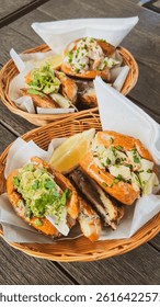 Assorted bread rolls featuring American lobster and prawn rolls with avocado, cilantro, lettuce, and spring onions, alongside a melted cheese and thin ham bread. Served on parchment paper in a basket.
