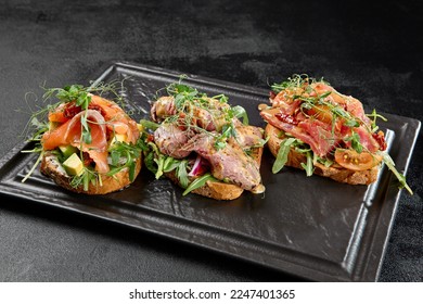Assorted bruschetta in black concrete background. Antipasti set: tuna, salmon and roastbeef bruschetta on stone plate. Fish bruschetta and meat bruschetta in minimal style