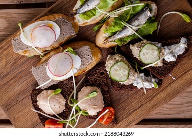 Assorted Bruschetta With Rabbit Liver, Cod Liver, Tulle On Lettuce And Cucumber Slices. On Top Of The Bruschetta Are Pea Microgreen Sprouts. Bruschetta Lie On A Wooden Board, The Board Stands On A Woo