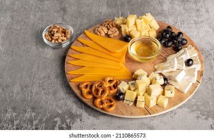 Assorted Cheese On A Wooden Plate On A Gray Background With A Copy Space. Wine Appetizer With Honey, Grapes, Pretzels And Walnuts On A Round Platter. Horizontal Orientation, No People, Copy Space