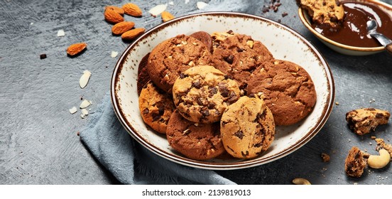Assorted chocolate chip cookies on wooden plate with cup of coffee on dark background, top view, panorama.