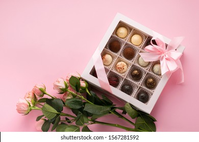 Assorted Chocolates In A Box Decorated With Pink Satin Ribbon, Bow Of Fresh Rose Flowers On A Pink Background. Top View