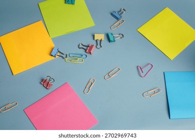 Assorted colorful sticky notes, binder clips, and paperclips scattered on a blue desk surface. Creative workspace concept with vibrant stationery elements arranged randomly.