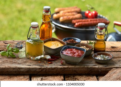 Assorted condiments and spices for a summer barbecue on a rustic wood picnic table outdoors in the garden with sausages grilling over a fire behind