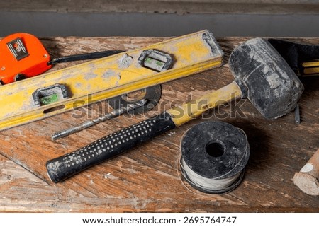 Assorted construction tools on worn wooden surface, including spirit level, rubber mallet, drill bit, twine, and tape measure in realistic DIY workspace setup.