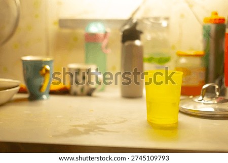 Assorted cups on dusty table, student study break with colorful plastic cup, ceramic mugs, water bottle, subtle water rings and fingerprints, soft window glow, relaxed communal atmosphere,