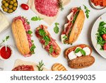 Assorted delicious baguette sandwiches filled with thinly sliced ham or salami and fresh green lettuce or arugula for breakfast arranged on white background, top view with copy space