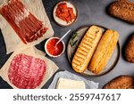 Assorted delicious ingredients for baguette sandwiches filled with thinly sliced ham or salami or Salchichon extra or jamon for breakfast arranged on dark background, top view with copy space