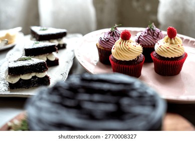 Assorted desserts on a table, featuring chocolate cake slices and red velvet cupcakes with cream frosting and raspberries.