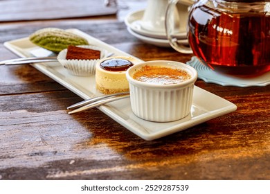 Assorted desserts with tea on a rustic table. A beautifully arranged assortment of desserts on a rectangular white plate, featuring crème brûlée, cheesecake, matcha madeleine, and a chocolate truffle.