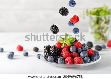 Assorted different berries with mint leaves on a glass plate on white marble table against light grey wall. Blueberry, blackberry, raspberry  mix group fying falling 