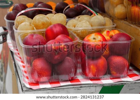 Assorted fresh fruits in clear containers at a market, including red apples and yellow plums, arranged on a checkered tablecloth