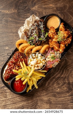 Assorted fried snacks including onion rings, chicken wings, ribs with cheese, fries, popcorn, sauces and salad, served on a black plate on dark wooden table.