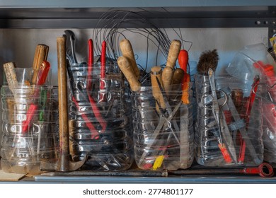 Assorted hand tools including hammers pliers screwdrivers and metal implements stored in reused plastic containers on a workshop shelf showing organized storage and practical workspace setup