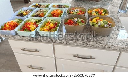 Assorted healthy salads are packed in rectangular containers on a granite kitchen counter. Ingredients include greens, carrots, cucumbers, and peppers.