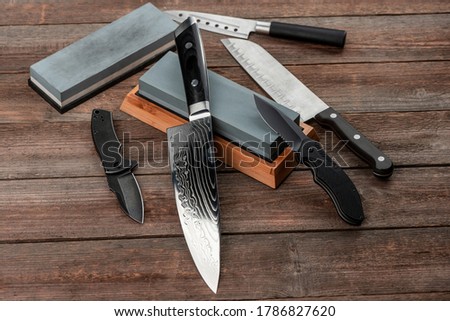 Assorted knives and sharpening stones on a rustic wooden table, top view. Jackknife, cheese knife, Santoku and chef's knife.