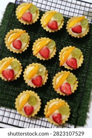 Assorted mini fruit tarts with custard cream and fresh fruit topping arranged neatly on cooling rack over green textured surface.