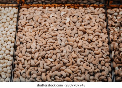 Assorted nuts including cashews, hazelnuts, and macadamia nuts are neatly arranged in clear containers at a market, showcasing a variety of textures and colors