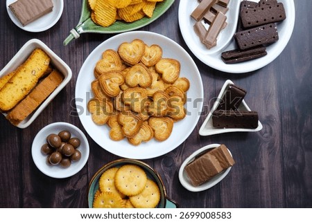 Assorted party snacks on table featuring chocolate cream sandwich biscuits, sugar coated heart cookies, wafer chocolates, chocolate coated caramel balls, salted crackers, and crispy potato chips.