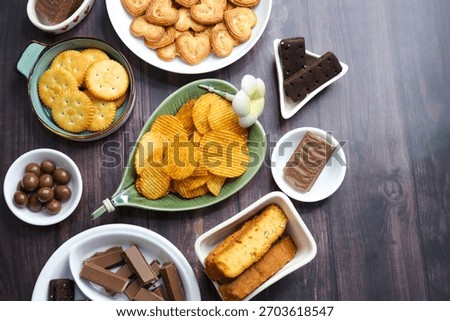 Assorted party snacks on table featuring chocolate cream sandwich biscuits, sugar coated heart cookies, wafer chocolates, chocolate coated caramel balls, salted crackers, and crispy potato chips.