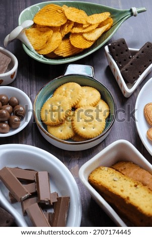 Assorted party snacks on table featuring chocolate cream sandwich biscuits, sugar coated heart cookies, wafer chocolates, chocolate coated caramel balls, salted crackers, and crispy potato chips.