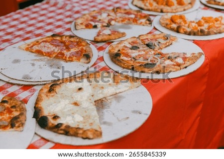 Assorted pizzas are arranged on round boards, showcasing different toppings at a lively outdoor food fair.