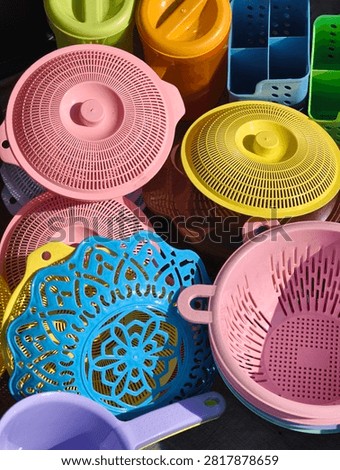 Assorted plastic containers, strainers, and home accessories in red, green, blue, pink, and yellow colors arranged outdoors under direct sunlight.