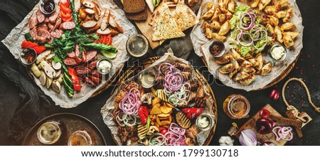 Assorted snacks for beer in plates, delicious grilled meat, vegetables, fries potatoes, smoked sausages, bbq sauce, chips, bread, pita and beer in glass on dark background, top view, toning