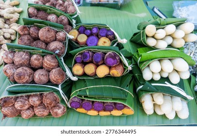 Assorted Steamed Root Vegetables Wrapped in Banana Leaves at Thai Street Food Market. Traditional Thai snack featuring steamed taro, purple sweet potato, and cassava neatly wrapped in banana leaves.