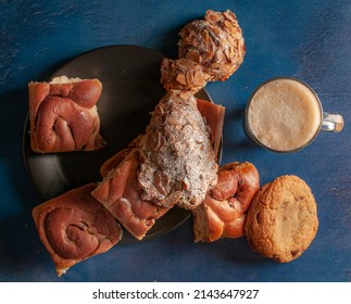 Assorted Sweet Bread On A Dark Plate With A Hot Cup Of Coffee Cocoa Chocolate Moka Cappuccino On The Side, On A Blue Kitchen Table, Horizontal Flat Lay Overhead Or Top View Shot.