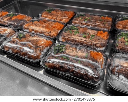 Assorted takeout bento boxes displayed in a refrigerated case. Each container features rice, meat, and vegetables neatly packed and sealed with clear plastic lids for convenient grab and go meals.