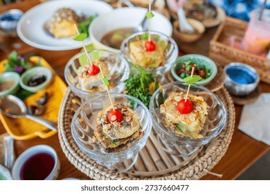 Assorted Thai appetizers served in small glass cups on a traditional tray with many side dishes in the background, photographed in natural light.