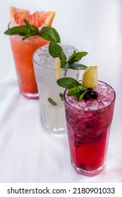 Assorted Three Types Of Lemonade, Classic Lemonade, Grapefruit Lemonade And Blackcurrant Lemonade. Drinks In Tall Glass Glasses With Ice, Lime Slices And Mint Leaves. The Dishes Are On A White Tablecl