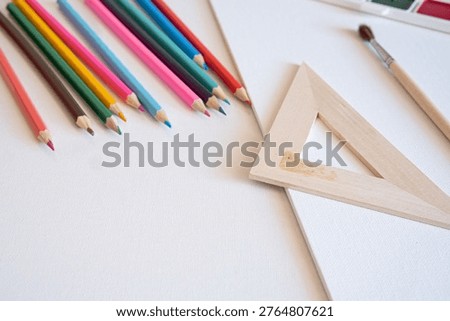 Assorted wooden pencils, drafting triangle, paintbrush, and blank canvas arranged on a white surface, representing the preparatory stage of analog artistic creation