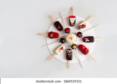 An Assortment Of Cakesicles And Cake Pop For Christmas, Featuring Santa, Reindeer Stars Christmas Tree On A White Plate In A Circle