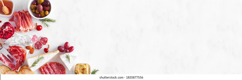 Assortment Of Cheese And Meat Appetizers. Overhead View Corner Border On A White Marble Banner Background With Copy Space.