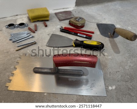 Assortment of Construction Tools and Materials Including Notched Trowel, Screws, and Measuring Tape on a Concrete Surface