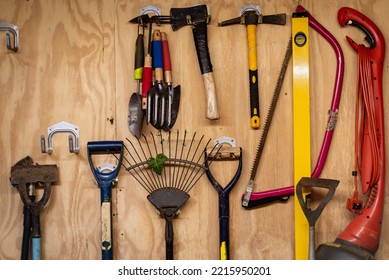 Assortment of DIY gardening tools and equipment hanging organised on wooden wall inside garden shed. Tools include rake, shovel, hammer, fork, trowel, spirit level measure, saw, axe, hatchet etc. - Shutterstock ID 2215950201