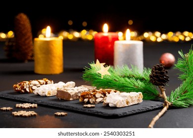 Assortment of gourmet nougat and nut bars on black slate plate under holiday ambiance with candles and evergreen sprigs, pinecone golden star with warm blurred lights perfect for celebrating Christmas - Powered by Shutterstock