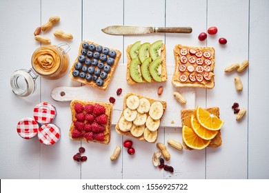 Assortment of healthy fresh breakfast toasts. Bread slices with peanut butter and various fruits and ingredients on side. Placed on white wooden table. Top view, with copy space.