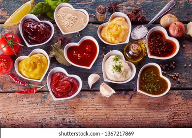 Assortment of marinades, sauces and dressings in individual heart shaped bowls surrounded by scattered spices, herbs and condiments on rustic wood